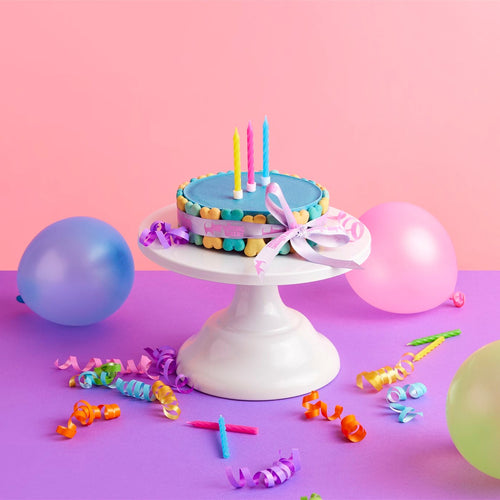 A round birthday cake with blue frosting and colorful candles sits on a white pedestal stand, surrounded by pastel balloons, party streamers, and ribbons on a pink and purple background.