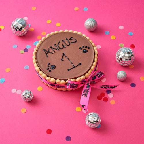 A round chocolate cake or cookie decorated with a pink ribbon, placed on a bright pink background with scattered confetti and silver disco ball ornaments. The top is brown and personalized with the name 'ANGUS' and the number '1'.